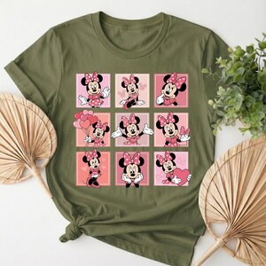 May include: Khaki green t-shirt with a grid of nine Minnie Mouse illustrations. Each square shows Minnie in different poses, wearing a pink polka-dot bow and dress. The background squares alternate between pink and peach tones.