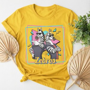 May include: Mustard yellow t-shirt featuring a graphic of two zebras wearing sunglasses. The zebras are in front of a geometric design with the word "Zebros" in a stylized font. The shirt has short sleeves and a crew neck.