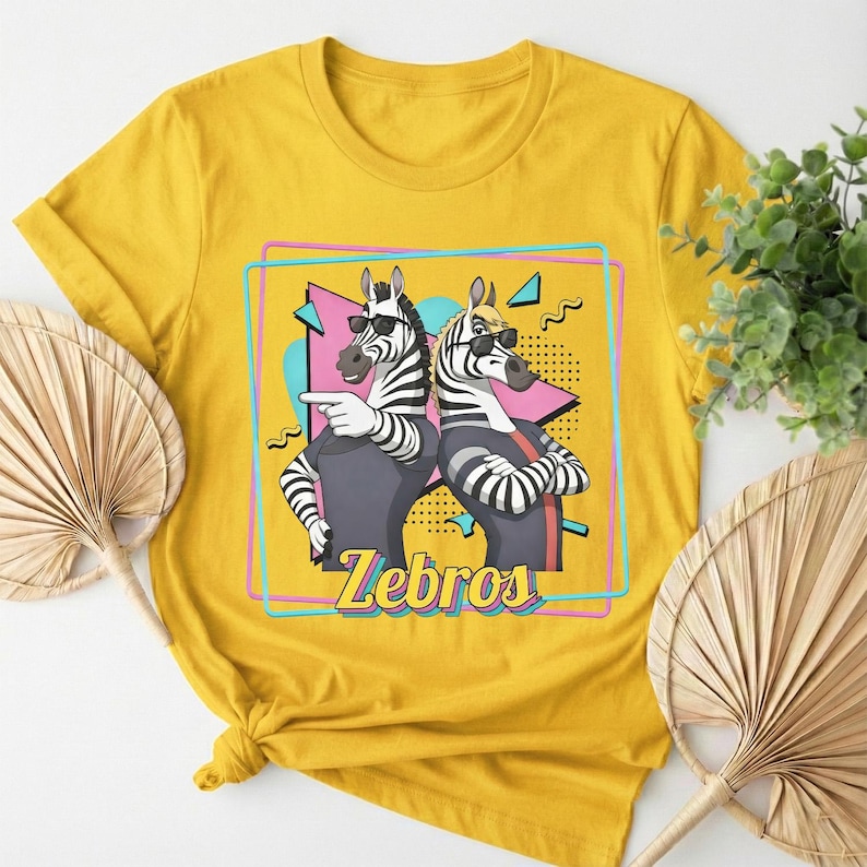 May include: Mustard yellow t-shirt featuring a graphic of two zebras wearing sunglasses. The zebras are in front of a geometric design with the word "Zebros" in a stylized font. The shirt has short sleeves and a crew neck.