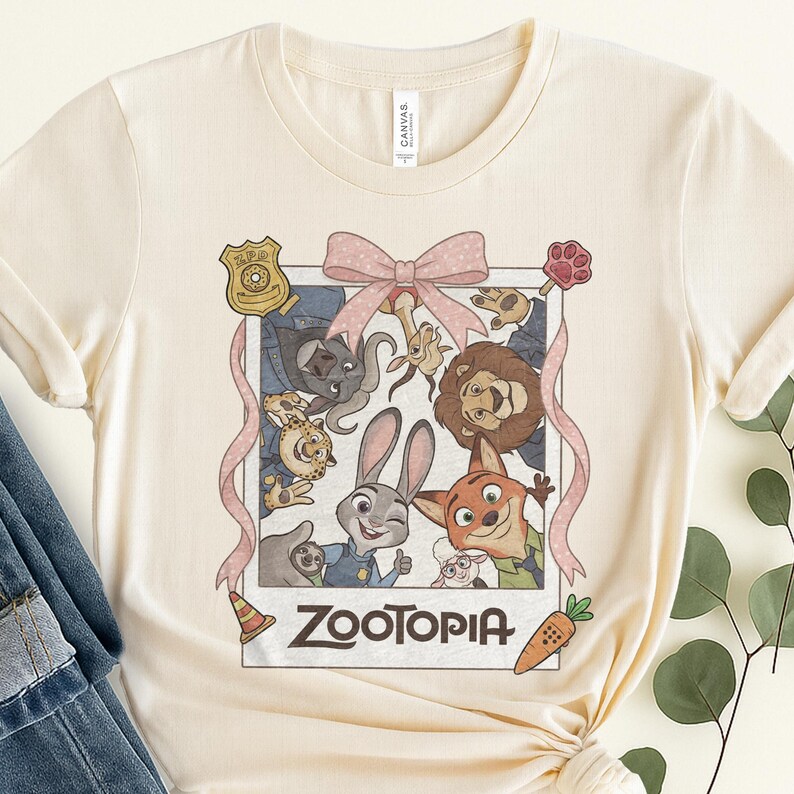 K&ouml;nnte beinhalten: Cremefarbenes T-Shirt mit einer Grafik aus dem Animationsfilm Zootopia. Das Design zeigt Charaktere wie Judy Hopps und Nick Wilde, umrahmt von einer rosa Schleife und einem Band. Ein Polizeiausweis, ein Pfotenabdruck und eine Karotte sind ebenfalls enthalten.