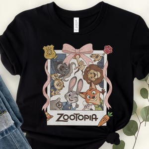 K&ouml;nnte beinhalten: Schwarzes T-Shirt mit einer gerahmten Grafik von Charakteren aus dem Animationsfilm "Zoomania". Das Bild zeigt Judy Hopps, Nick Wilde und andere Charaktere, mit einer rosa Schleife und dem Filmtitel unten. Eine Karotte und ein Pfotenabdruck sind ebenfalls enthalten.