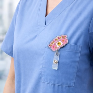 May include: A pink badge reel with the text "Certified Boo-Boo Fixer" and a smiling face on a bandage design. The reel is attached to a clear plastic holder and is clipped to a blue scrub top pocket.