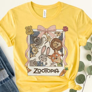 K&ouml;nnte beinhalten: Gelbes T-Shirt mit einem Zootopia-Motiv. Das Design zeigt Charaktere wie Judy Hopps und Nick Wilde, mit dem Wort "Zootopia" unten. Eine rosa Schleife und B&auml;nder umrahmen das Bild.