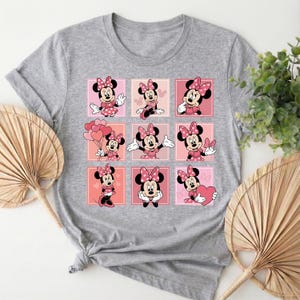 May include: A grey t-shirt with a grid of nine images of Minnie Mouse. Each image shows Minnie in different poses, set against a pink background. Minnie Mouse is wearing a pink dress and bow. The t-shirt is made of a soft material.