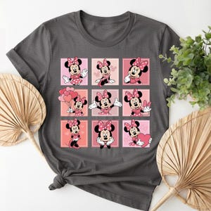May include: Dark grey t-shirt with a grid design of Minnie Mouse illustrations. Each square features Minnie in a pink dress and bow, set against a gradient pink background. The design is a playful and colourful graphic.