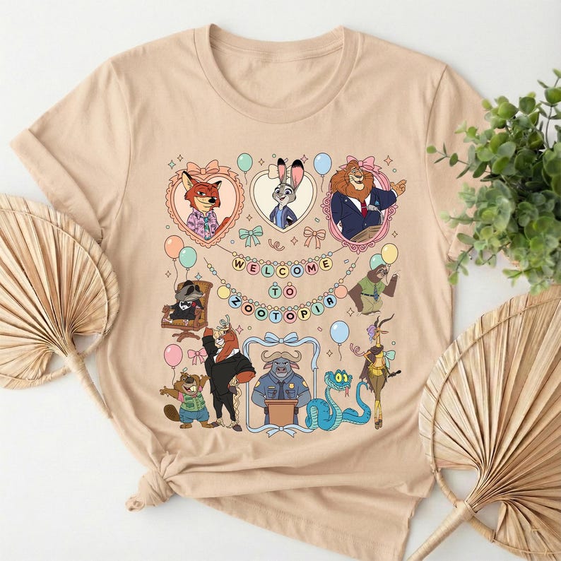 Puede incluir: Camiseta beige con un gr&aacute;fico colorido de personajes de la pel&iacute;cula animada Zootopia. El dise&ntilde;o incluye el texto "WELCOME TO ZOOTOPIA" y varios personajes de animales en marcos en forma de coraz&oacute;n, globos y elementos decorativos.