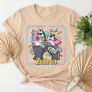 May include: Beige t-shirt featuring a graphic of two zebras wearing sunglasses, with the word "Zebros" in yellow. The zebras are in a retro 80s style, with a pink and blue square border and a heart shape in the background.