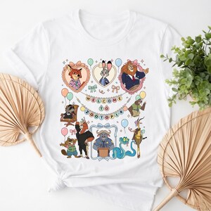 Puede incluir: Camiseta blanca con un gr&aacute;fico colorido de personajes de Zootopia, incluyendo un zorro, un conejo y un le&oacute;n, con el texto "WELCOME TO ZOOTOPIA". El dise&ntilde;o incluye globos y marcos en forma de coraz&oacute;n.