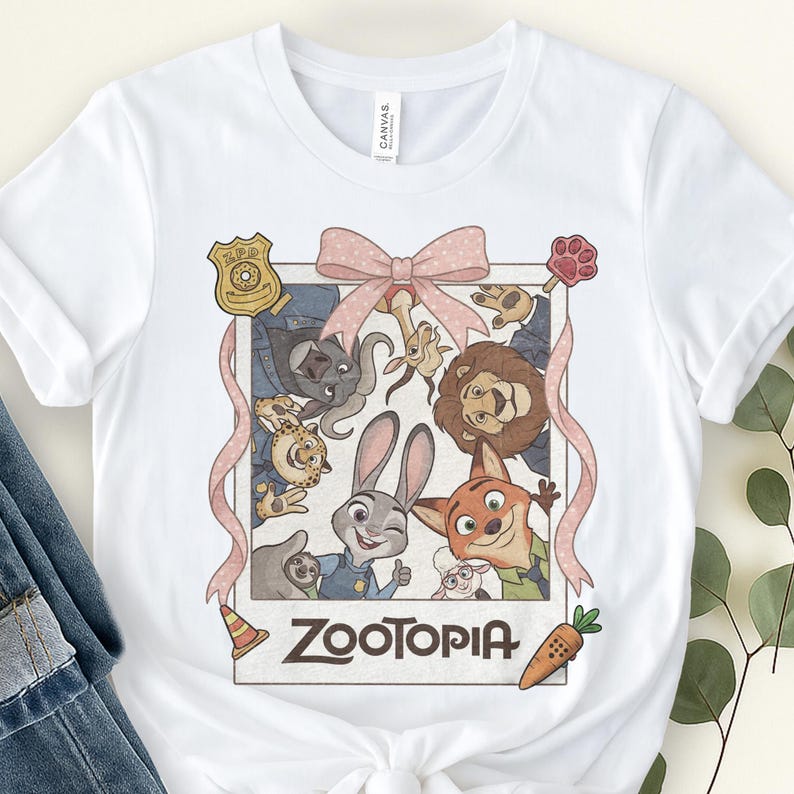 K&ouml;nnte beinhalten: Wei&szlig;es T-Shirt mit einer gerahmten Grafik von Charakteren aus dem Animationsfilm "Zoomania". Das Design umfasst Judy Hopps, Nick Wilde und andere Charaktere, mit einer rosa Schleife und dem Wort "Zoomania" unten. Eine Karotte und ein Pfotenabdruck sind ebenfalls enthalten.