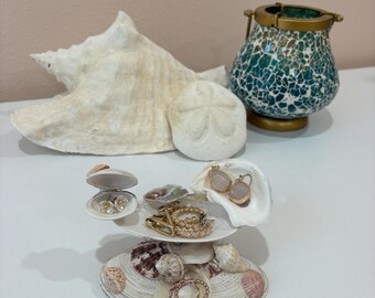 May include: A seashell jewelry holder with multiple levels, displaying various earrings, rings, and necklaces. The holder is made of seashells, with a large conch shell and a teal mosaic candle holder in the background. The jewelry is gold and rose gold.