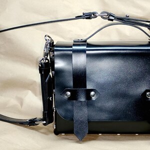 May include: Black leather satchel bag with a top handle and adjustable shoulder strap. The bag features silver-tone hardware, including clasps and rivets. Vertical straps with buckle closures secure the front flap.