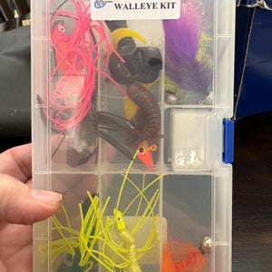 Puede incluir: Una caja de aparejos de plástico transparente etiquetada como "WALLEYE KIT" contiene varios señuelos de pesca. Los señuelos son de diferentes colores, incluyendo rosa, amarillo, naranja y morado. Algunos señuelos tienen una apariencia plumosa, mientras que otros son más sólidos.