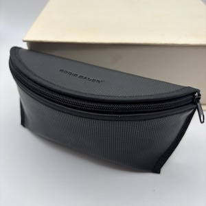 May include: A black, semi-circular zippered pouch with a textured, perforated surface. The brand name Eddie Bauer is embossed in black lettering on the top flap. The pouch features a single black zipper closure with a metal pull tab.