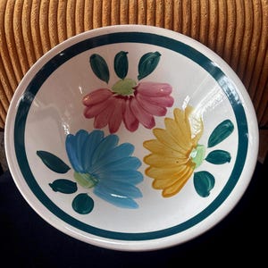 May include: White ceramic bowl with a teal rim and floral design. The bowl features three large flowers in pink, blue, and yellow, with green leaves. The bowl is round and has a simple, yet colorful design.