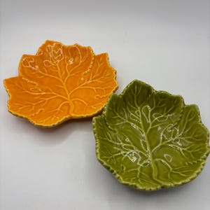 May include: Two ceramic dishes shaped like leaves, one in a vibrant orange and the other in a deep olive green. Both dishes feature a glossy finish and detailed, embossed vein patterns that mimic the natural structure of a real leaf.