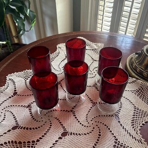 May include: A set of five drinking glasses featuring deep red glass bowls and clear glass bases. The glasses have a cylindrical shape with a smooth finish and are arranged in a cluster on a surface, showcasing the contrast between the red and clear glass.