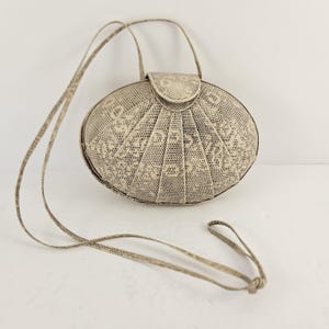 May include: A beige, oval-shaped handbag with a textured, patterned surface. The bag features a flap closure and a long, thin strap. The design includes a short handle and a longer, adjustable strap for shoulder or crossbody wear. The bag's overall aesthetic is vintage.