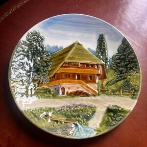 May include: A round ceramic decorative plate featuring a raised relief scene of a brown wooden house with a thatched roof. The landscape includes green trees, a wooden fence, a dirt path, and a small blue stream flowing in the foreground.