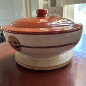 May include: A round ceramic serving bowl with a glossy, solid reddish-brown lid featuring a central knob. The bowl has a speckled off-white base with a horizontal dark brown band and circular accents, resting on a circular light wood base.