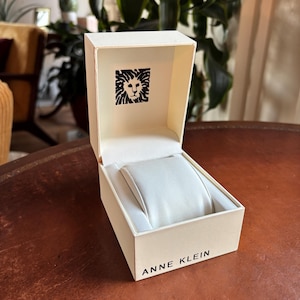 May include: An open, off-white Anne Klein watch box with a white interior and a watch pillow. The box features the Anne Klein logo on the inside lid and the brand name printed on the front.