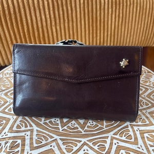 May include: A dark brown leather wallet with a flap closure. The wallet has a decorative gold-colored embellishment on the front. The wallet is rectangular and appears to be made of a smooth leather material.