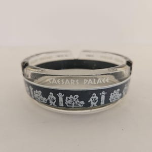 May include: A clear glass ashtray featuring the text CAESARS PALACE in white lettering. The ashtray has a black band around the center decorated with a repeating white graphic pattern of figures and columns in a classical style.
