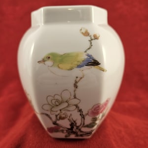 May include: White ceramic vase with a hexagonal top and a rounded body. The vase features a painted bird with green, yellow, and blue plumage perched on a branch with flowers. The flowers are white and pink, with brown stems.