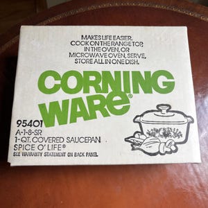 May include: A white cardboard box for a Corning Ware 1-qt. covered saucepan in the Spice O' Life pattern. The box features green text, black line art of the cookware, and the printed text: Makes life easier. Cook on the range top, in the oven, or microwave oven, serve, store all in one dish.