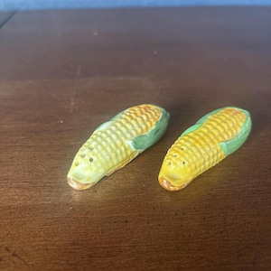 May include: A pair of ceramic salt and pepper shakers shaped like ears of corn. The shakers are yellow with green husks and small holes for dispensing. The set is designed to resemble fresh corn on the cob.