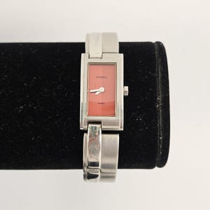 May include: A rectangular Fossil watch with a salmon-colored face and silver-tone steel band. The watch face displays the Fossil logo and the word "STEEL". The watch is displayed on a black surface.
