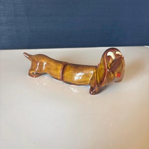 May include: A ceramic dachshund dog figurine in shades of brown with a glossy finish. The dog has white accents around the eyes and a red mouth. The figurine is a decorative item.