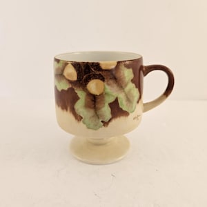 May include: A white ceramic mug with a black handle and the text "But First, Coffee" in a bold, black font. The mug is set against a blurred background, suggesting a cozy setting.