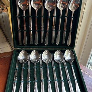 May include: A set of ten silver-toned metal spoons and ten matching pairs of chopsticks arranged in a dark green velvet-lined storage case. Each spoon features a smooth, reflective bowl and a slender handle with a subtle, embossed swirl design.