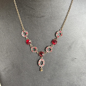 May include: A vintage-style necklace with a gold-tone chain and pendant. The necklace features a series of red gemstone accents and small clear crystals. The pendant has a teardrop shape with a small gold-tone bell at the bottom.