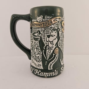 May include: Dark green ceramic beer stein with a black handle. The stein features a white and black illustration of a bear in a tuxedo, with the text "Hamm's 1974" and "Grand Marshall".