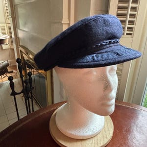 May include: A dark navy blue wool-blend fiddler cap featuring a structured crown and a short, stiff brim. The hat is adorned with a decorative braided cord trim across the front and an intricate embossed floral pattern on the brim surface.