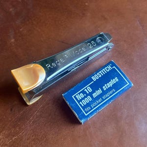 May include: A silver Regal Model 25 stapler with an orange handle next to a blue box of Bostitch No. 10 mini staples. The box reads "1000 mini staples" and "fits pocket staplers".