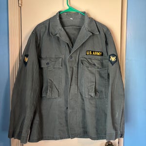 May include: An olive green U.S. Army jacket with two chest pockets and two lower pockets. The jacket has a button-up front and a collar. Patches with a gold emblem are on the sleeves. The text "U.S. ARMY" is above the right chest pocket.