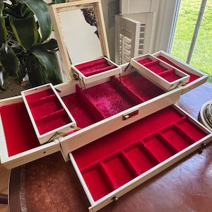 May include: An open, vintage jewelry box with a cream-colored exterior and a bright red velvet interior. The box has multiple compartments, drawers, and a small mirror. The box is on a brown table.
