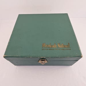 May include: A vintage, rectangular, teal-colored box with a gold-colored clasp and the words "Merchants National" printed in gold lettering. The text "THE FULL SERVICE BANK OF CEDAR RAPIDS" is printed below the main text.