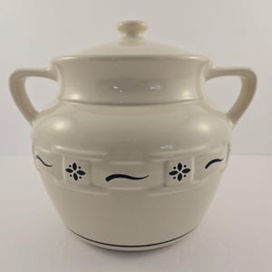 May include: Cream-colored ceramic jar with a lid and two handles. The jar features a decorative band with dark blue floral and wave patterns. A dark blue line encircles the base of the jar. The jar is suitable for kitchen storage.