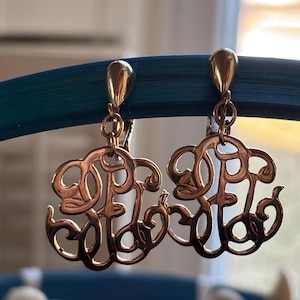 May include: A pair of gold-toned drop earrings. Each earring features an ornate, stylized design with a looping, cursive monogram. The earrings have a teardrop-shaped top and a hinged closure. The earrings are displayed against a blurred background.