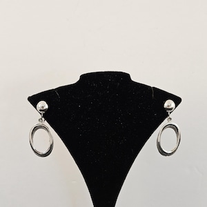 May include: Silver-toned oval drop earrings. Each earring features a small, round stud at the top, connected to a larger, open oval shape. The earrings are displayed on a black jewelry stand against a white background.
