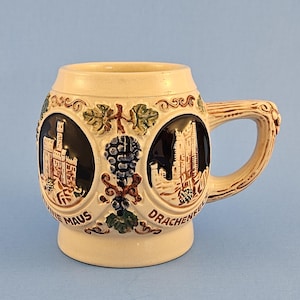 May include: A cream-colored ceramic beer mug featuring two dark oval medallions with raised relief depictions of castles. The text Ruine Maus and Drachenfels is printed below the medallions, accented by green leaves and blue grape clusters.