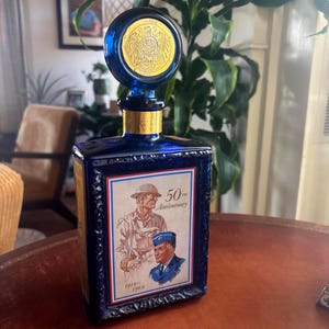 May include: A cobalt blue glass decanter with a round stopper featuring a gold emblem. The decanter has a rectangular body with a framed image of two men and the text "50th Anniversary 1919-1969".