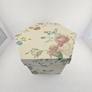 May include: A hexagonal box with a floral pattern featuring pink, blue, and white roses on a cream background. The box has a lid and sides, and the floral design is repeated on all surfaces. The box is likely for storage or decorative purposes.