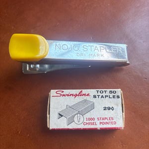 May include: A vintage silver stapler with a yellow handle, labeled "NO.10. STAPLER DRI MARK", next to a small box of "Swingline" staples. The box is white with red text and a diagram of staples.