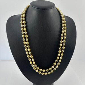 May include: A gold-toned necklace with two strands of round beads. The necklace is displayed on a black mannequin bust against a white background. The beads have a matte finish, and the necklace is likely made of metal.