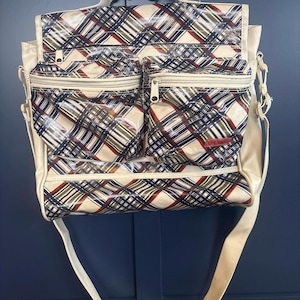 May include: Cream-colored messenger bag with a plaid pattern in red, navy, and olive green. The bag has two front zippered pockets and a long shoulder strap. The flap and pockets feature a glossy finish.