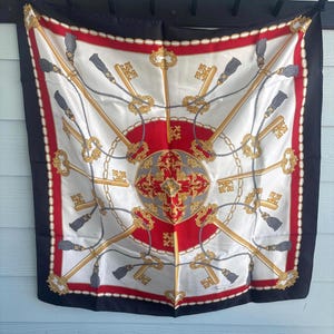 May include: A square silk scarf featuring a white center with a red circular medallion, decorated with gold keys, grey tassels, and rope motifs. The border is red with a white chain pattern, finished with a wide black edge on all four sides.